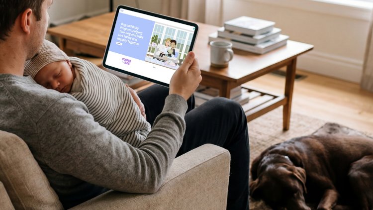 Over the shoulder view of a man sitting on a couch with a newborn baby, holding a tablet to view the Cooper and Kids online program, Dog and Baby Program: Helping Your Dog and Baby Live Safely and Happily Together, with a brown labrador sleeping at his feet.