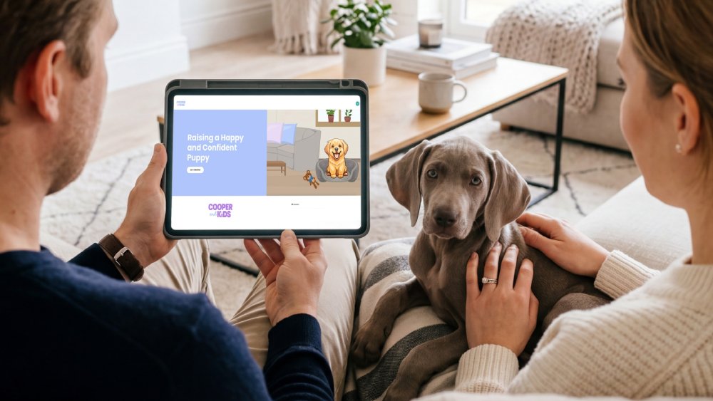 A couple sit on a couch with their new puppy, looking at a tablet displaying the Cooper and Kids online training program, Raising a Happy and Confident Puppy.
